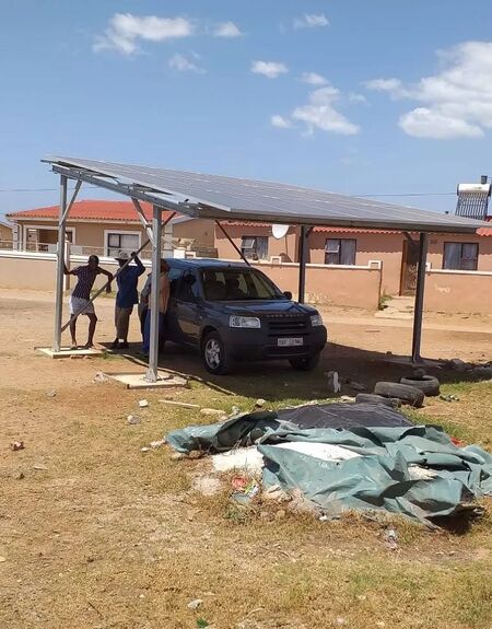 Picture of two people standing next to an SUV beneath a carport outfitted with 15 solar PV panels. According to the original publisher, the panels generated 5 kilowatts of electricity that was fed into the KwaZakhele Township's municipal grid.
