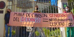 Three protestors stand in front of a gate and hold a banner reading, in all capitals, "Mina de carbón del Descanso = Genocidio, neocolonialismo, ecocidio."